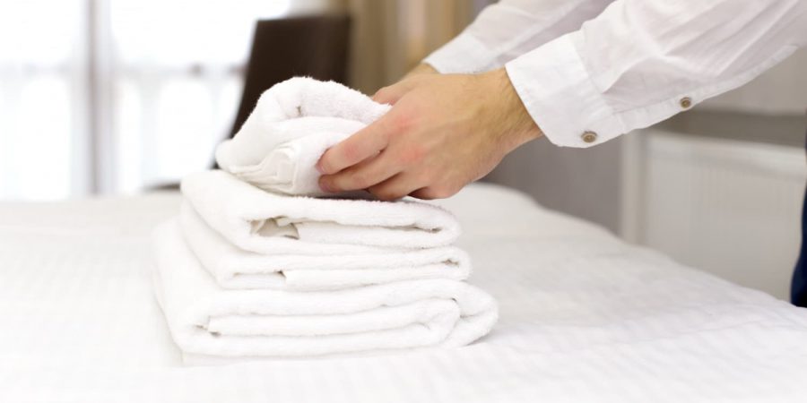 Person legt frische weiße Handtücher auf ein Hotelbett, während ein Reinigungsdienst für Hotels das Zimmer vorbereitet.