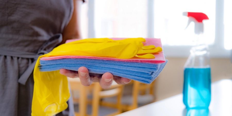 Person hält Putztücher und gelbe Handschuhe in der Hand, daneben steht eine Sprühflasche für die Unterhaltsreinigung.